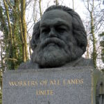 The gravestone of Karl Marx in North London, England. The gravestone is surrounded by green trees. The gravestone is made of marble and has a bust of Marx carved atop it. Inscribed in gold below the bust are the words "WORKERS OF ALL LANDS UNITE."